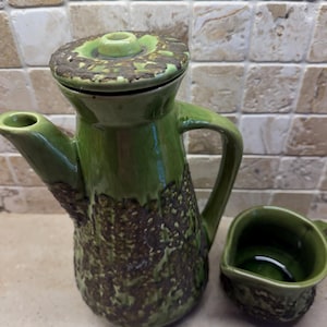 May include: A green ceramic coffee pot and creamer set. The coffee pot features a textured, dark brown pattern on the lower section and a lid with a small knob. The creamer is a small, open container with a handle. The set is on a light-coloured surface.