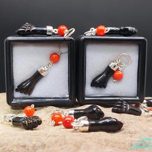 Puede incluir: Una colección de dijes con forma de mano negra con detalles de metal plateado y cuentas esféricas de color naranja brillante. Algunos dijes se muestran dentro de dos cajas cuadradas negras con insertos blancos, otros están esparcidos.