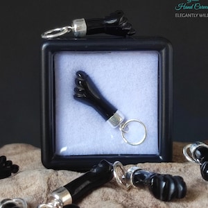 May include: Black onyx hand carved charm in the shape of a fist. The charm is attached to a silver chain and ring. The charm is displayed in a black box with a white interior. The text "Gaia Hand Carved Elegantly Wild!" is visible in the top right corner of the image.