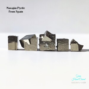 May include: Five cube-shaped Navajún pyrite mineral specimens from Spain. The metallic pyrite crystals are arranged in a row against a white background. The text "Navajún Pyrite From Spain" is at the top, and "Gaia Hand Carved, Elegantly Wild!" is at the bottom.