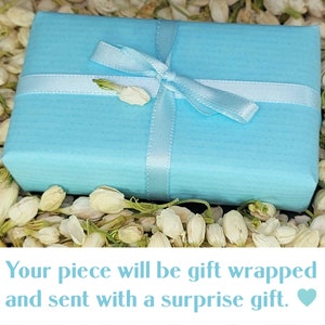 May include: A light blue gift box tied with a white ribbon and surrounded by white flowers. The text on the image reads "Your piece will be gift wrapped and sent with a surprise gift."