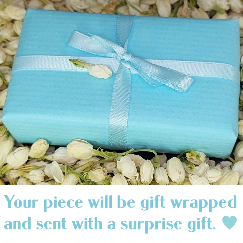 May include: A light blue gift box tied with a white ribbon and surrounded by white flowers. The text on the image reads "Your piece will be gift wrapped and sent with a surprise gift."