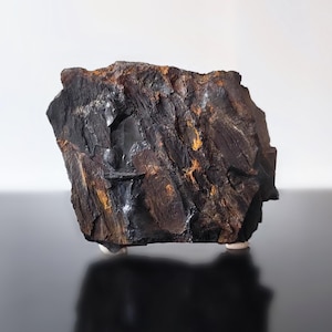 May include: A dark, jagged rock specimen with a rough, uneven surface texture. The stone features deep charcoal and black tones with prominent patches of rusty orange and brown mineral deposits embedded within its crevices and layered structure.