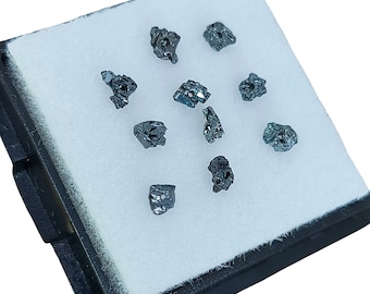 May include: A collection of ten small, dark grey, rough, and irregularly shaped crystals are displayed in a white, velvet-lined, black box.