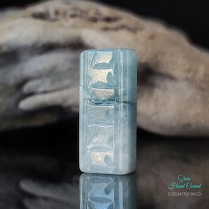 May include: A hand-carved jade pendant with a light blue color and intricate designs. The pendant is rectangular in shape and has a smooth, polished finish. The text "Gaia Hand Carved" and "ELEGANTLY WILD!" is visible below the pendant.