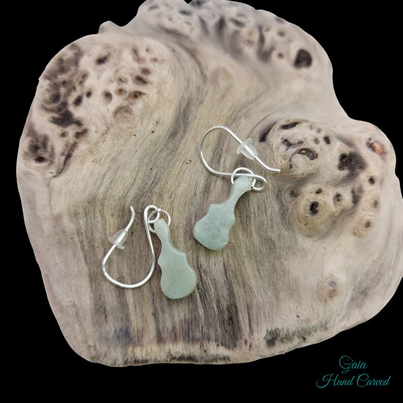 May include: A pair of silver dangle earrings with green jade guitar pendants. The earrings are displayed on a piece of driftwood. The text "Gaia Hand Carved" is visible in the bottom right corner of the image.