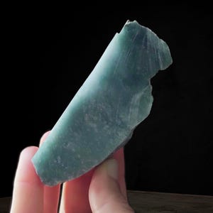 May include: A hand holding a piece of raw, teal-colored jade. The stone has a smooth, polished surface with visible natural striations and a rough, uneven edge. The jade is set against a dark background.