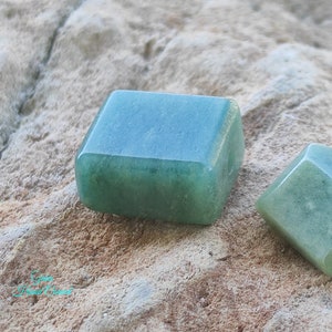 May include: Close-up of two polished, square-shaped green aventurine stones. The stones have rounded edges and a slightly translucent appearance. The background is a textured, light brown surface.
