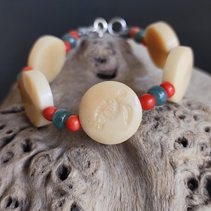 May include: Cream-colored bracelet featuring a turtle design. The bracelet has large, oval beads and a central round bead with a turtle carving. Red and teal beads accent the design, with a silver clasp. A unique piece of jewelry.