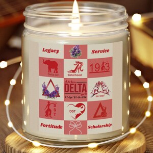 Puede incluir: Una vela encendida en un tarro de cristal transparente con una etiqueta con gráficos y texto en rosa y blanco. La etiqueta incluye las palabras "Legacy", "Service" y "1943". La vela está sobre un posavasos de madera, con una guitarra y un libro en el fondo.