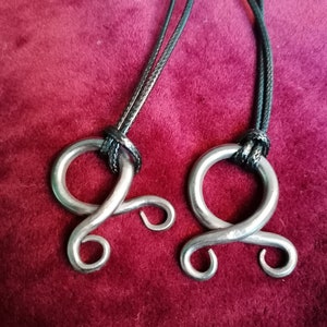 May include: Two silver metal pendants with a unique design, each hanging from a black cord. The pendants are shaped like a circle with two loops extending from the top.