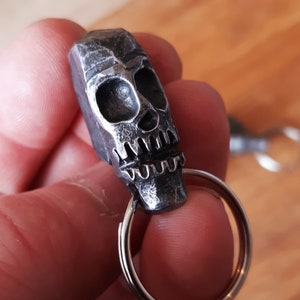 May include: A silver-coloured skull-shaped keyring. The skull has detailed eye sockets, a nose cavity, and a jagged jawline. The keyring is attached to a silver split ring. The metal has a textured, hammered appearance.