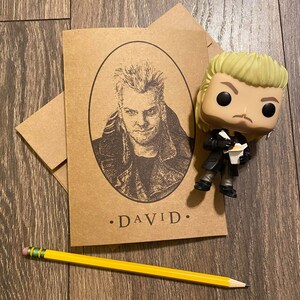 May include: A brown card with a black and white illustration of a man with spiky hair. The card has the text "DAVID." printed on it. A yellow pencil is lying on a wooden surface next to the card. A small, yellow-haired figurine of the man is standing next to the card.