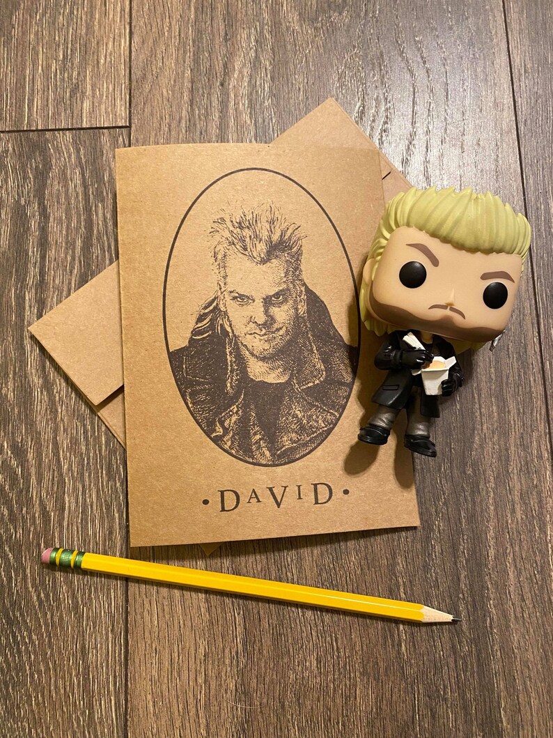 May include: A brown card with a black and white illustration of a man with spiky hair. The card has the text "DAVID." printed on it. A yellow pencil is lying on a wooden surface next to the card. A small, yellow-haired figurine of the man is standing next to the card.