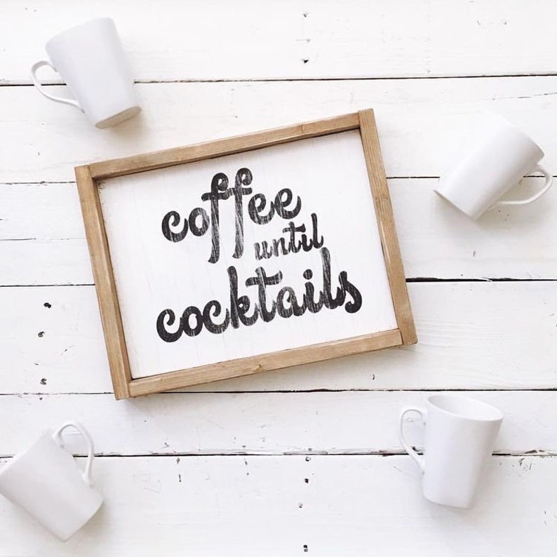 May include: A wooden sign with the text "coffee until cocktails" in black script font. The sign is sitting on a white wooden surface with four white mugs.