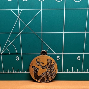 May include: A round, bronze-colored pendant featuring a detailed engraving of a man on a camel holding a scorpion. The pendant has a small loop at the top for hanging. The background is a green cutting mat with white grid lines.