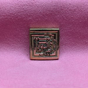 Può includere: Una spilla vintage quadrata color oro con un design a rilievo dettagliato. Il rilievo raffigura figure in stile classico, forse una scena religiosa o storica. La spilla è posta su uno sfondo rosa.