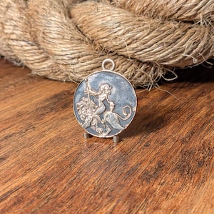 May include: A silver-toned pendant with a detailed relief of a woman holding a spear, riding a winged creature. The pendant is round with a dark blue background and a small loop at the top. The pendant is displayed on a wooden surface.