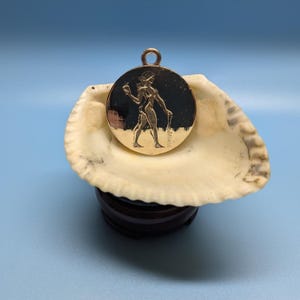 May include: A gold-coloured pendant featuring a raised image of a woman holding a pitcher. The pendant is resting on a white seashell.