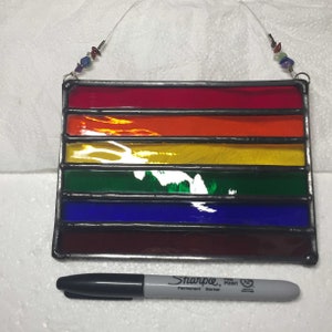 May include: A stained glass rainbow flag hanging from a string with beads. The flag is made of six horizontal stripes in the colours red, orange, yellow, green, blue, and purple.