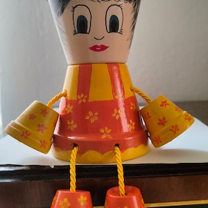 Könnte beinhalten: Handbemalte Blumentopfpuppe aus Terrakotta. Die Puppe hat ein Gesicht mit blauen Augen, roten Lippen und schwarzem Haar. Der Körper ist orange und gelb mit floralen Akzenten, Arme und Beine sind kleine Töpfe, die mit gelbem Seil befestigt sind.