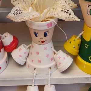 Pode incluir: Uma boneca de vaso de terracota artesanal com um corpo branco, detalhes florais rosa e um adorno de cabeça de renda e fita. A boneca tem olhos azuis pintados, um sorriso vermelho e braços e pernas de vaso pendurados. A boneca está sentada em uma prateleira branca.