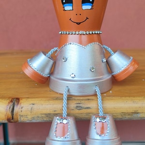 Puede incluir: Una figura caprichosa de maceta de arcilla hecha a mano con una cara sonriente, adornada con pintura plateada y pedrería. La figura tiene brazos y piernas hechas de macetas más pequeñas, conectadas con cuerda. La figura está sentada sobre una superficie de madera.