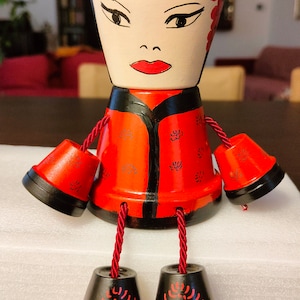Könnte beinhalten: Dekorative, handbemalte Terrakotta-Topfpuppe. Die Puppe hat ein bemaltes Gesicht, rote Lippen und einen schwarzen Hut mit rosa Blumen. Sie trägt eine rote Jacke mit schwarzem Besatz und hat hängende Arme und Beine aus kleinen Töpfen und rotem Seil.