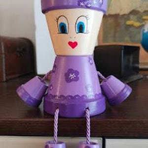 Peut inclure: Un pot en terre cuite peint à la main en forme de figurine, avec un visage beige clair, des yeux bleus et une bouche en forme de cœur rouge. La figurine porte une robe et un chapeau violets, ornés de motifs floraux. Le pot contient des plantes vertes et a des pieds violets suspendus.