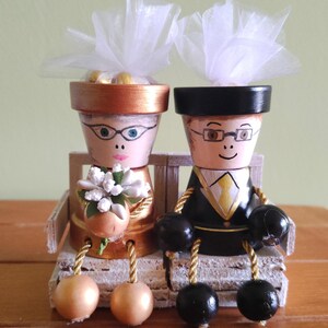 May include: Hand-painted wooden bride and groom figurines. The bride wears glasses and holds a bouquet. The groom is in a tuxedo. Both are seated on a small bench. Each figure has a flower pot head and dangling ball feet.