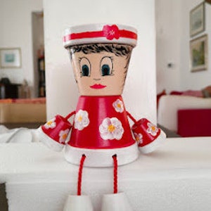 Pode incluir: Boneca artesanal feita de vasos de terracota pintados. A boneca apresenta um vestido vermelho com detalhes florais brancos, um chapéu branco com uma faixa vermelha e sapatos vermelhos e brancos. A boneca tem um rosto pintado com uma expressão sorridente.