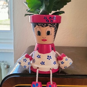 Pode incluir: Vaso de flores de terracota pintado à mão em forma de boneca. A boneca tem um vestido rosa e branco com motivos florais azuis, cabelo preto e um rosto pintado. Contém uma planta verde com flores cor-de-rosa.