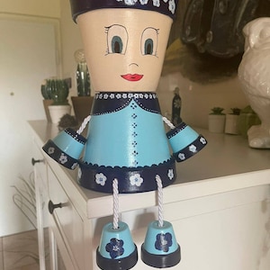 Op de afbeelding: Een handbeschilderde terracotta bloempot in de vorm van een pop. De pot heeft een lichtblauwe jurk, donkerblauwe accenten en een lachend gezicht. Witte touwbenen hangen naar beneden en de hoed is donkerblauw met witte bloemaccenten.
