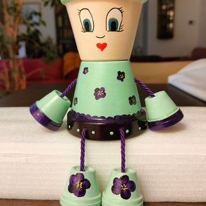 Op de afbeelding: Handbeschilderde bloempotpop met een lichtgroen lichaam, paarse bloemen, een lachend gezicht en paarse touwarmen en -benen. De pop is ontworpen als decoratie.