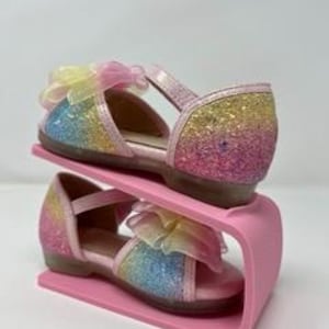 May include: A pair of pink glittery sandals with rainbow ombre and a yellow bow. The sandals are on a pink shoe stand.