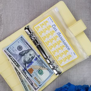 May include: A yellow binder with a clear plastic pocket containing a yellow and white sticker that says "Car Insurance" with a grid of 30 circles. The binder has a silver ring mechanism and is open to reveal a stack of US dollar bills.