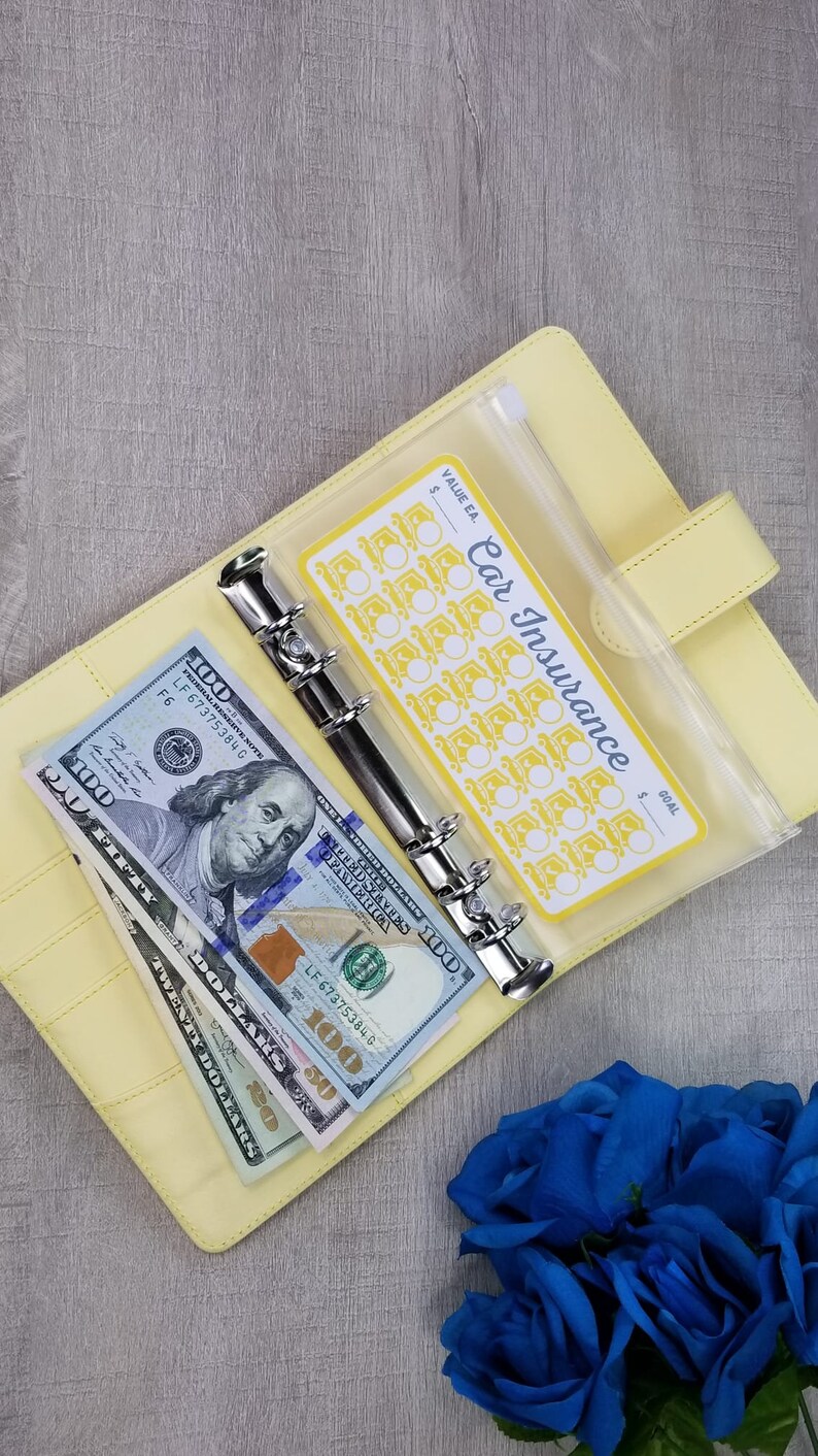 May include: A yellow binder with a clear plastic pocket containing a yellow and white sticker that says "Car Insurance" with a grid of 30 circles. The binder has a silver ring mechanism and is open to reveal a stack of US dollar bills.