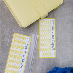 May include: Two clear plastic zippered pouches with yellow car icons and the text "Car Insurance" and "Car Note Payoff" with a yellow planner and blue roses on a gray wood background.