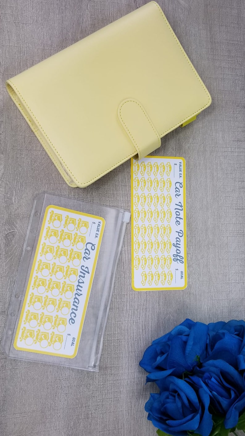 May include: Two clear plastic zippered pouches with yellow car icons and the text "Car Insurance" and "Car Note Payoff" with a yellow planner and blue roses on a gray wood background.