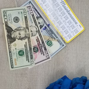 May include: A clear plastic pouch with a zipper closure containing a sheet of car maintenance stickers and three US dollar bills: a $20, a $50, and a $100 bill. The pouch is on a light brown surface with a bouquet of blue roses in the lower right corner.