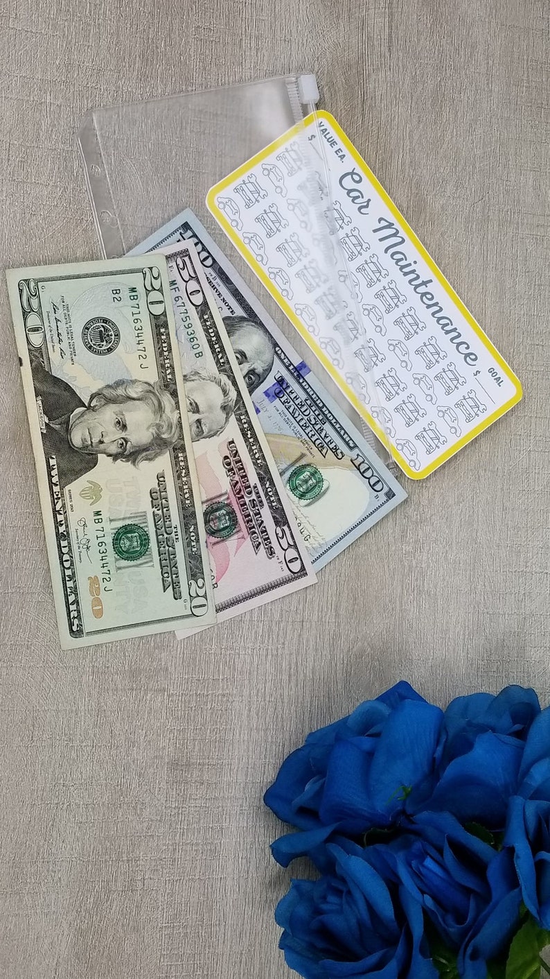 May include: A clear plastic pouch with a zipper closure containing a sheet of car maintenance stickers and three US dollar bills: a $20, a $50, and a $100 bill. The pouch is on a light brown surface with a bouquet of blue roses in the lower right corner.