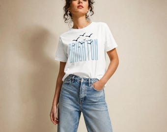 Könnte beinhalten: Ein weißes Kurzarm-T-Shirt mit dem Wort BRIGHTON in großen, hellblauen vertikalen Buchstaben, ergänzt durch eine kleine Grafik aus dunkelblauen fliegenden Vögeln über dem Text, kombiniert mit einer hellblauen Jeanshose.