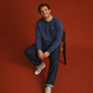 Pode incluir: Uma camisola de manga comprida azul-marinho com gola redonda e um pequeno logótipo bordado no peito, combinada com calças de ganga azul-escuras e ténis de lona bege-claros. A peça apresenta um design simples com punhos canelados.