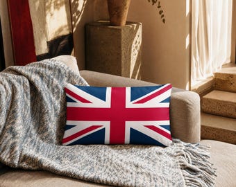 May include: A rectangular throw pillow featuring the Union Jack flag design in red, white, and blue. The pillow is positioned on a textured gray throw blanket that is draped over a sofa, showcasing the bold geometric pattern of the flag.