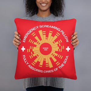 Puede incluir: Almohada roja con el texto "Emergency Screaming Pillow" y un gráfico de una onda sonora con el texto "Scream Here" en el centro. El texto "Fully Absorbs Cries of the Soul" está escrito alrededor del gráfico de la onda sonora.