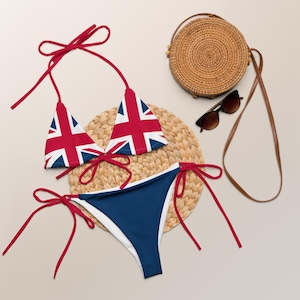 May include: A two-piece bikini set featuring the Union Jack flag design. The top has triangle cups with red ties, and the bottom is navy blue with white trim and red ties. A woven bag, sunglasses, and a woven mat are also in the image.