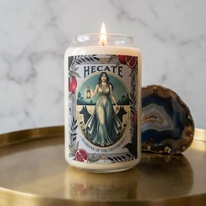 Könnte beinhalten: Ein brennendes, klares Glas-Kerzenglas mit einem Etikett mit dem Wort "HECATE" über einer Illustration einer Frau, die eine Laterne hält, flankiert von zwei schwarzen Hunden. Der Text "GUARDIAN OF THE CROSSROADS" steht ebenfalls auf dem Etikett.
