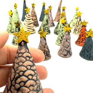 May include: A collection of ceramic Christmas trees in various colors and patterns. Each tree is topped with a yellow star. The trees have textured surfaces and are displayed against a white background. Some trees are held in a hand.