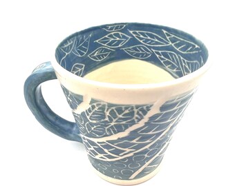 May include: A handmade ceramic mug with a blue exterior and a white interior. The mug features a leaf pattern in white, with a matching blue handle. The mug is ideal for coffee, tea, or other beverages.