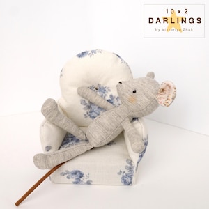 May include: A grey stuffed mouse toy with a pink-patterned ear rests on a small armchair. The armchair is upholstered in white fabric with blue floral patterns. A brown stick is in the foreground. The text "10 x 2 DARLINGS by Viktoriya Zhuk" is in the upper right corner.